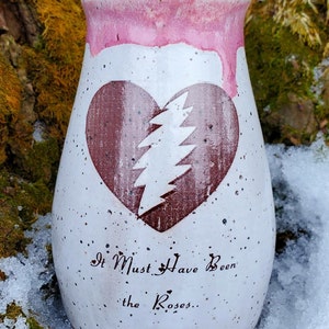 May include: A speckled white ceramic vase with a pink rim and a heart design. The heart is a dark red color with a white lightning bolt design. The text "It Must Have Been the Roses..." is written below the heart.