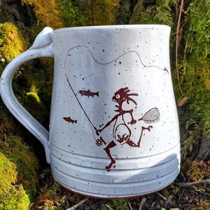 May include: White ceramic mug with a brown illustration of a stick figure fishing. The illustration includes a fishing rod, a net, and three fish. The mug has a speckled glaze.