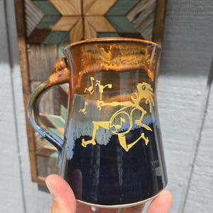 Puede incluir: Una taza de cerámica con base azul oscuro, que transiciona a un borde marrón y dorado. La taza presenta una ilustración dorada de un músico tocando un instrumento. El asa es una mezcla de azul y marrón.