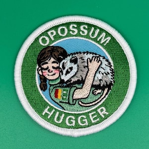 May include: Embroidered patch featuring a cartoon illustration of a person hugging an opossum. The patch has a green border with the words "OPOSSUM HUGGER" in white. The background is blue.