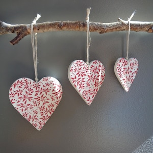 May include: Three white heart-shaped ornaments with a red floral pattern hanging from a natural wooden branch by thin twine strings. The hearts vary in size, with the largest on the left and the smallest on the right, all featuring a repeating botanical print.