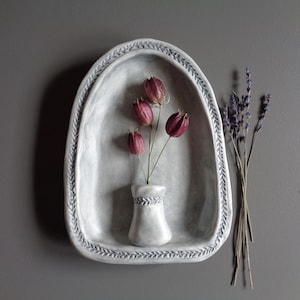 May include: A gray ceramic wall shelf with a decorative border holds a small vase with three dark pink flowers. A bundle of dried lavender is to the right. The shelf is mounted on a gray wall.