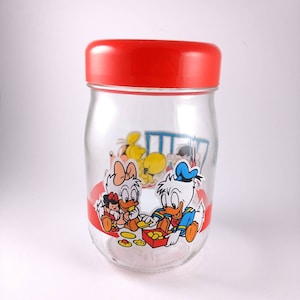 May include: A glass jar with a red lid and a cartoon image of Donald Duck and Daisy Duck with a baby duck. The jar has a red stripe around the middle and a red stripe around the top.