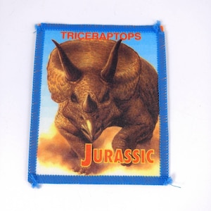 Op de afbeelding: Een blauwe en oranje geborduurde patch met een bruine Triceratops-dinosaurusillustratie en de tekst "TRICERAPTOPS JURASSIC".