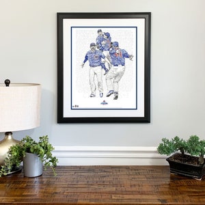 May include: Framed artwork featuring a celebratory baseball team in blue uniforms. The image is a detailed illustration composed of text, with a black frame and white matting. The artwork is displayed on a light gray wall.