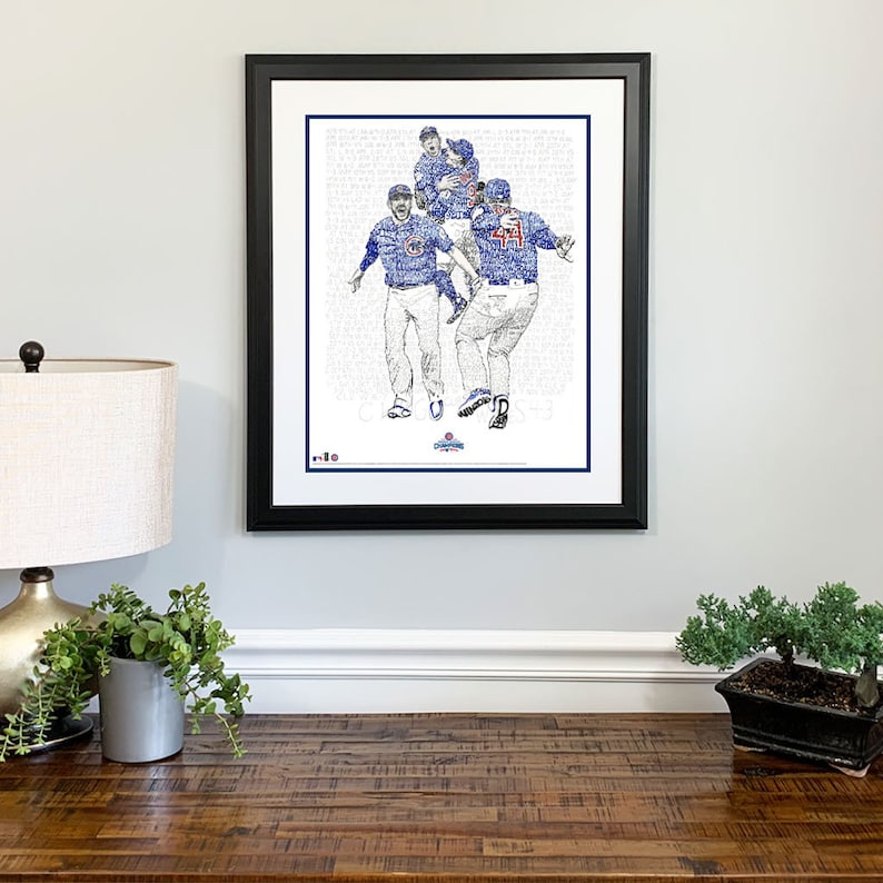 May include: Framed artwork featuring a celebratory baseball team in blue uniforms. The image is a detailed illustration composed of text, with a black frame and white matting. The artwork is displayed on a light gray wall.