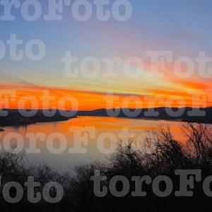 May include: A scenic view of a river at sunset. The sky is a vibrant orange and pink, with a few clouds. The river is calm and reflective, and the trees on the far shore are silhouetted against the sky.