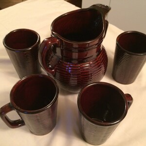 May include: A dark brown ceramic drinks set, comprising a pitcher and four cups. The pitcher has a handle and a wide spout. The cups include mugs with handles and tumblers. All pieces feature a ribbed texture.
