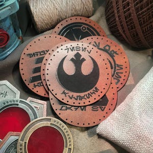 May include: A collection of leather patches and metal badges with a Star Wars theme. The patches are circular, with a brown leather appearance and black symbols and text. The metal badges have a red center and metallic borders. Also visible are spools of twine and a small cloth bag.