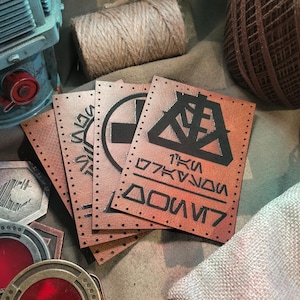 May include: Leather patches with black symbols and text are displayed. The patches are brown with stitched edges. A roll of twine and other metal objects are in the background. The text on the patch is in a stylized font.