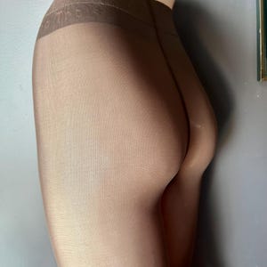 May include: Sheer, nude-colored tights. The waistband is a darker shade of brown with the word "Tights" printed on it. The tights are form-fitting and appear to be made of a smooth, stretchy material. The overall look is sleek and minimalist.