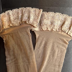 May include: A pair of sheer nude stockings featuring wide, decorative lace bands at the top. The lace trim has a detailed floral pattern with a scalloped edge, while the main body of the stockings is made of a fine, translucent, light tan fabric.