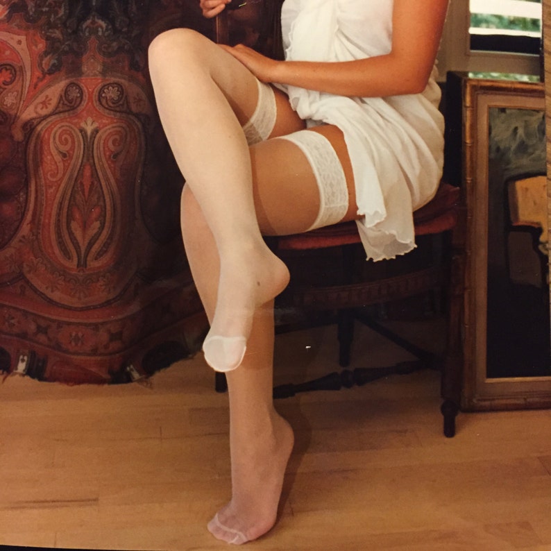 May include: Sheer white thigh-high stockings featuring a delicate lace band at the top. The hosiery is translucent, revealing the skin tone underneath, and is worn with a simple white garment that drapes over the upper legs while seated on a chair.