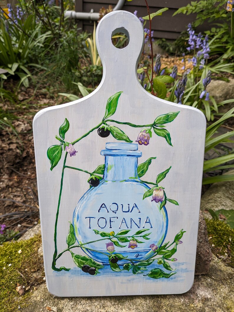 May include: A white wooden chopping board with a painted design of a blue glass bottle with the words "Aqua Tofana" on it. The bottle is surrounded by green leaves and purple flowers.