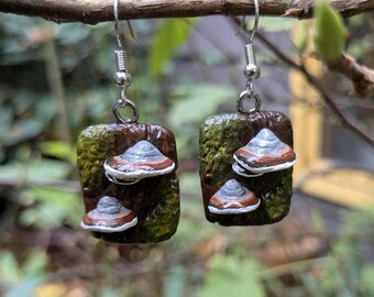 May include: Handmade earrings with a mushroom design. The earrings have a rectangular base with a textured, mossy green and brown appearance. Each earring has two or three mushroom-shaped accents with grey, white, and brown details, suspended from silver-coloured hooks.