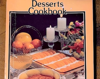 May include: A pink cookbook with the title "Desserts Cookbook" and the words "Favorite Recipes Press" on the bottom. The cover features a still life of a dessert in a white bowl with a blue and white floral pattern, surrounded by fruit, candles, and a silver serving dish with sliced peaches.