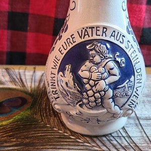 May include: A white ceramic pitcher with blue and white painted details. The pitcher features a scene of a man carrying grapes with the text "TRINKT WIE EURE VÄTER AUS STEIN DEN WEIN" around the scene.