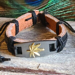 May include: A black leather bracelet with a silver charm featuring a gold marijuana leaf design. The bracelet has a black cord with a knot at the end.