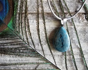 May include: A teal blue agate pendant necklace with a silver chain. The pendant is teardrop shaped and has a natural, organic pattern. The necklace is lying on a wooden surface with a peacock feather.
