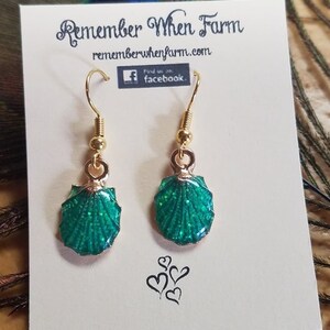 May include: A pair of gold earrings with green glitter seashell charms. The earrings are on a white card with the text "Remember When Farm" and a website address.