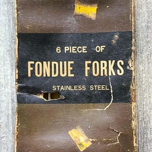May include: Vintage box of six stainless steel fondue forks. The box is brown with black lettering that reads "6 Piece of Fondue Forks Stainless Steel". The box has some wear and tear.