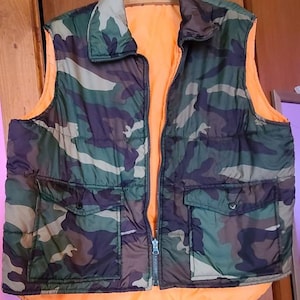 May include: A camouflage-patterned puffer vest with a bright orange lining. The vest has two zippered pockets and a zipper closure.