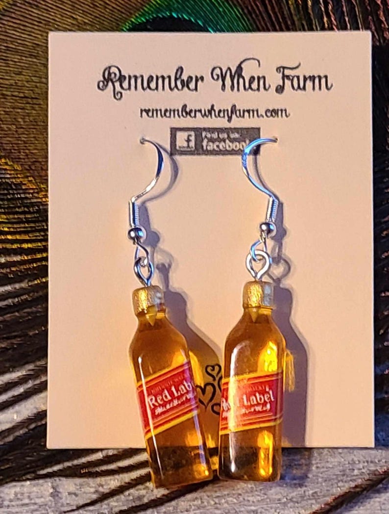 Puede incluir: Un par de pendientes de aro de plata con botellas de vidrio en miniatura con forma de botellas de whisky. Las botellas contienen un l&iacute;quido amarillo y tienen una etiqueta roja que dice "Red Label" con un borde dorado. Los pendientes est&aacute;n en una tarjeta blanca con el texto "Remember When Farm" y "rememberwhenfarm.com".