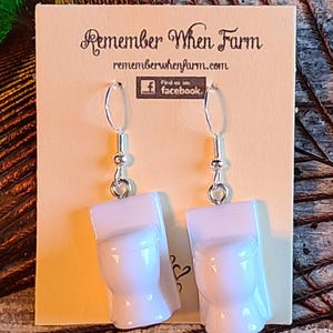 May include: White porcelain toilet earrings with silver hooks. The earrings are on a card that says "Remember When Farm" and "rememberwhenfarm.com".