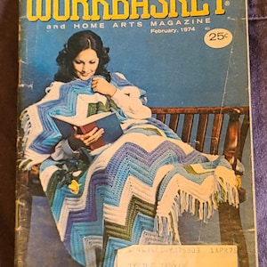 May include: The Workbasket and Home Arts Magazine, February 1974, features a woman reading while wrapped in a blue and white chevron patterned blanket. The magazine cover has a blue background with yellow text and a 25-cent price tag.