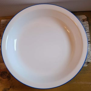 May include: A round, white enamelware pie dish or shallow bowl featuring a smooth, glossy finish and a distinct, thin navy blue rim that circles the entire outer edge of the vessel, providing a clean, high-contrast border to the white interior.