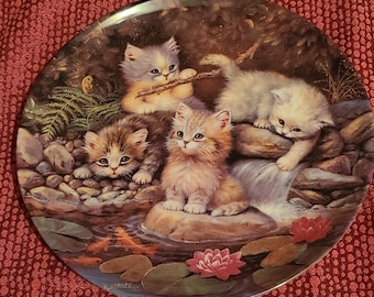 May include: A round ceramic plate with a woodland scene featuring four kittens playing by a stream. The kittens are white, brown, and gray with green foliage and pink flowers in the background. The plate is signed "J.Scholz".