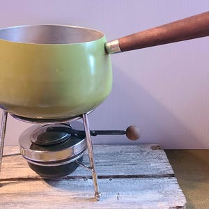 May include: A vintage fondue pot with a light green exterior and a wooden handle. The pot sits on a chrome stand with a burner below. The handle is a dark brown color. The pot is designed for cooking and serving fondue.