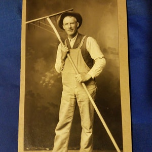May include: A black and white vintage photograph of a man holding a rake. The man is wearing a hat and overalls. The photo is labeled "Westerman."