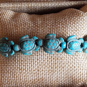 May include: A turquoise blue bracelet with a series of turtle charms. The bracelet is made of a dark string and the turtle charms are made of a smooth, polished stone.