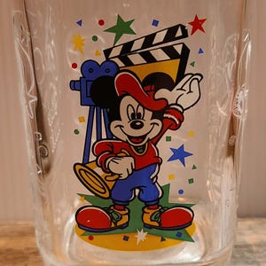 May include: A clear glass tumbler featuring a colorful graphic of Mickey Mouse wearing a red cap, red shirt, and blue shorts. Mickey holds a megaphone near a film clapperboard and camera. The glass includes the text 2000 and Disney branding.