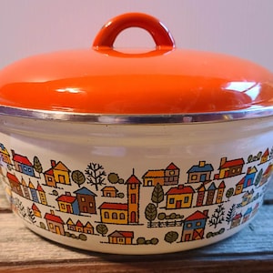 May include: A vintage enamelware pot with a bright orange lid and handle. The white pot features a colorful village scene with houses, trees, and a church. The pot has two side handles and a silver rim.