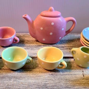 May include: A ceramic tea set featuring a pink teapot with white polka dots, four small cups in pink, blue, light green, and yellow, a matching creamer, and a stack of four small saucers in blue, yellow, pink, and light green with white dots.
