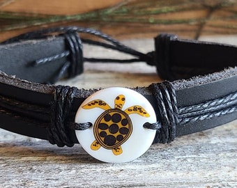 May include: Black leather bracelet featuring a white circular centerpiece with a yellow and brown turtle design. The bracelet has multiple black leather straps and adjustable black cords for closure. A casual accessory.