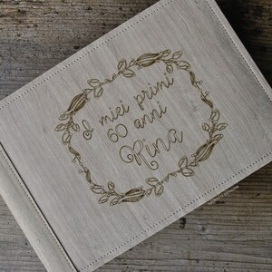 May include: A light grey wooden photo album featuring a floral wreath design and the Italian text "I miei primi 60 anni Rina". The album is closed and has a stitched border.