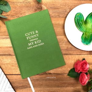 May include: A green journal with the text "Cute & Funny Things My Kid Says and Does" on the cover. The journal is on a wooden surface with a green cactus cookie, a bouquet of pink roses, and a white and gray striped towel.