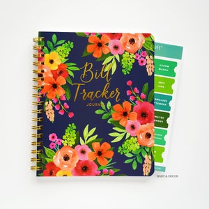 May include: A blue spiral bound journal with a floral design and gold lettering that reads "Bill Tracker Journal". The cover features a colorful floral pattern with red, orange, pink, and green flowers. A sticker sheet with monthly tabs is attached to the side of the journal.