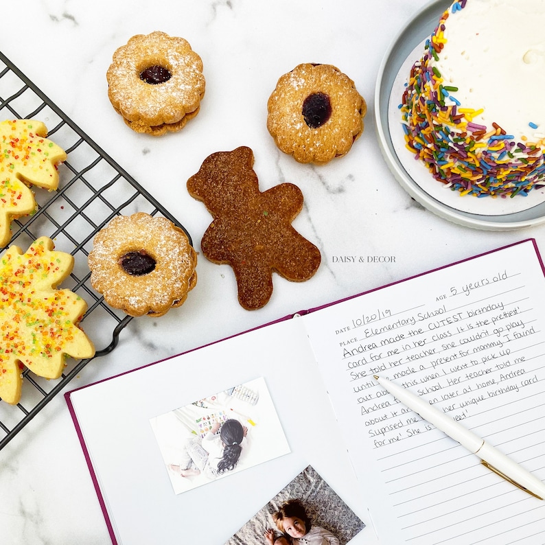 May include: A purple notebook with a handwritten note about a birthday celebration at an elementary school. The note mentions a birthday card made by a student named Andrea. The notebook is open to a page with a photo of a child and a pen on top of the page. There are also cookies and a cake with sprinkles on a white surface.