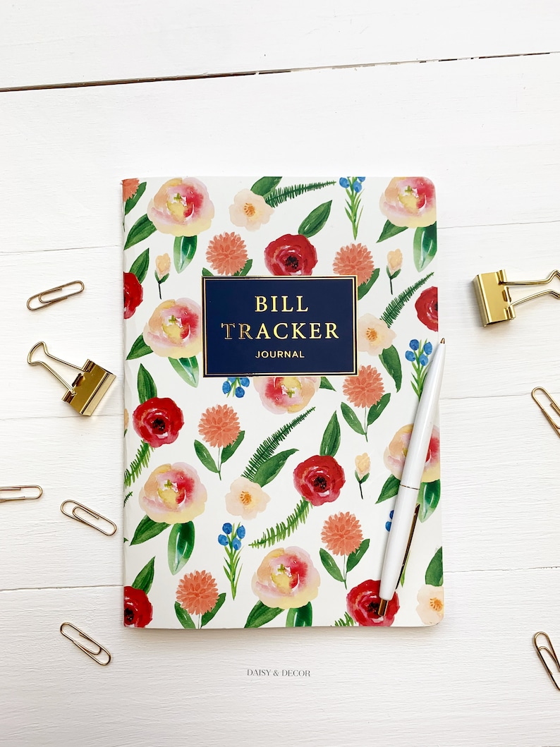 Puede incluir: Un cuaderno con estampado floral con el t&iacute;tulo "Bill Tracker Journal" en un recuadro azul marino. El cuaderno est&aacute; sobre una superficie de madera blanca con clips de papel dorados.