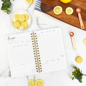 Op de afbeelding: Een wit receptenboek met een gouden spiraalbinding. Het boek heeft een gestreepte kaft met de woorden "Recipe" en "Daisy & Decor" erop gedrukt. Het boek is open op een pagina met koppen voor "Voorbereidingstijd", "Kooktijd", "Porties", "Ingredi&euml;nten", "Moeilijkheidsgraad", "Instructies", "Notities", "Herinneringen" en "Datum van de eerste bereiding".