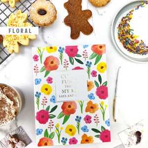 Puede incluir: Un diario blanco con un patr&oacute;n floral y el texto "Cute & Funny Things My Kid Says and Does" en la portada. El diario est&aacute; rodeado de galletas, un oso de peluche, un pastel y fotos.