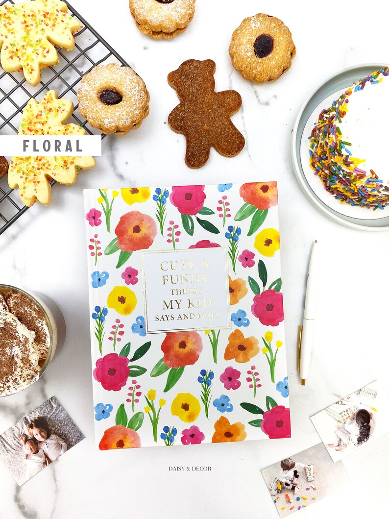 Puede incluir: Un diario blanco con un patr&oacute;n floral y el texto "Cute & Funny Things My Kid Says and Does" en la portada. El diario est&aacute; rodeado de galletas, un oso de peluche, un pastel y fotos.
