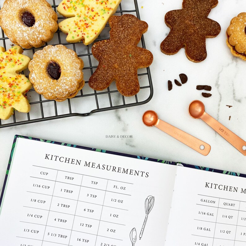May include: A close-up of a kitchen cookbook open to a page with a chart of kitchen measurements in cups, tablespoons, teaspoons, and fluid ounces. The chart is printed on a page with a dark blue background and a green floral pattern. Two copper measuring spoons are on a white surface next to the cookbook.