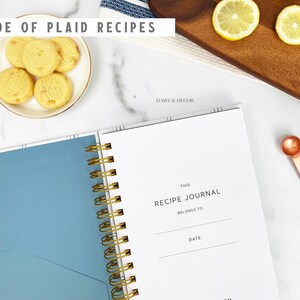 Op de afbeelding: Een wit receptenboek met een blauwe kaft en een gouden spiraalbinding. Het boek heeft de tekst "This Recipe Journal Belongs To" en "Date:" op de kaft.