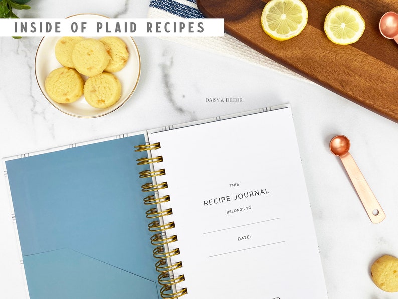 Op de afbeelding: Een wit receptenboek met een blauwe kaft en een gouden spiraalbinding. Het boek heeft de tekst "This Recipe Journal Belongs To" en "Date:" op de kaft.