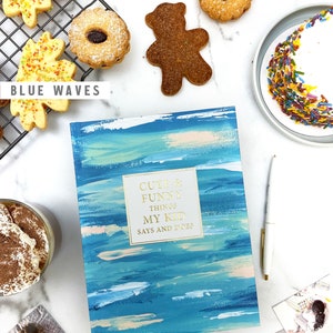 Puede incluir: Un libro azul y blanco con el t&iacute;tulo "Cute & Funny Things My Kid Says and Does" de Daisy & Decor. El libro est&aacute; rodeado de varios art&iacute;culos, que incluyen galletas, un oso de peluche y una taza de caf&eacute;.